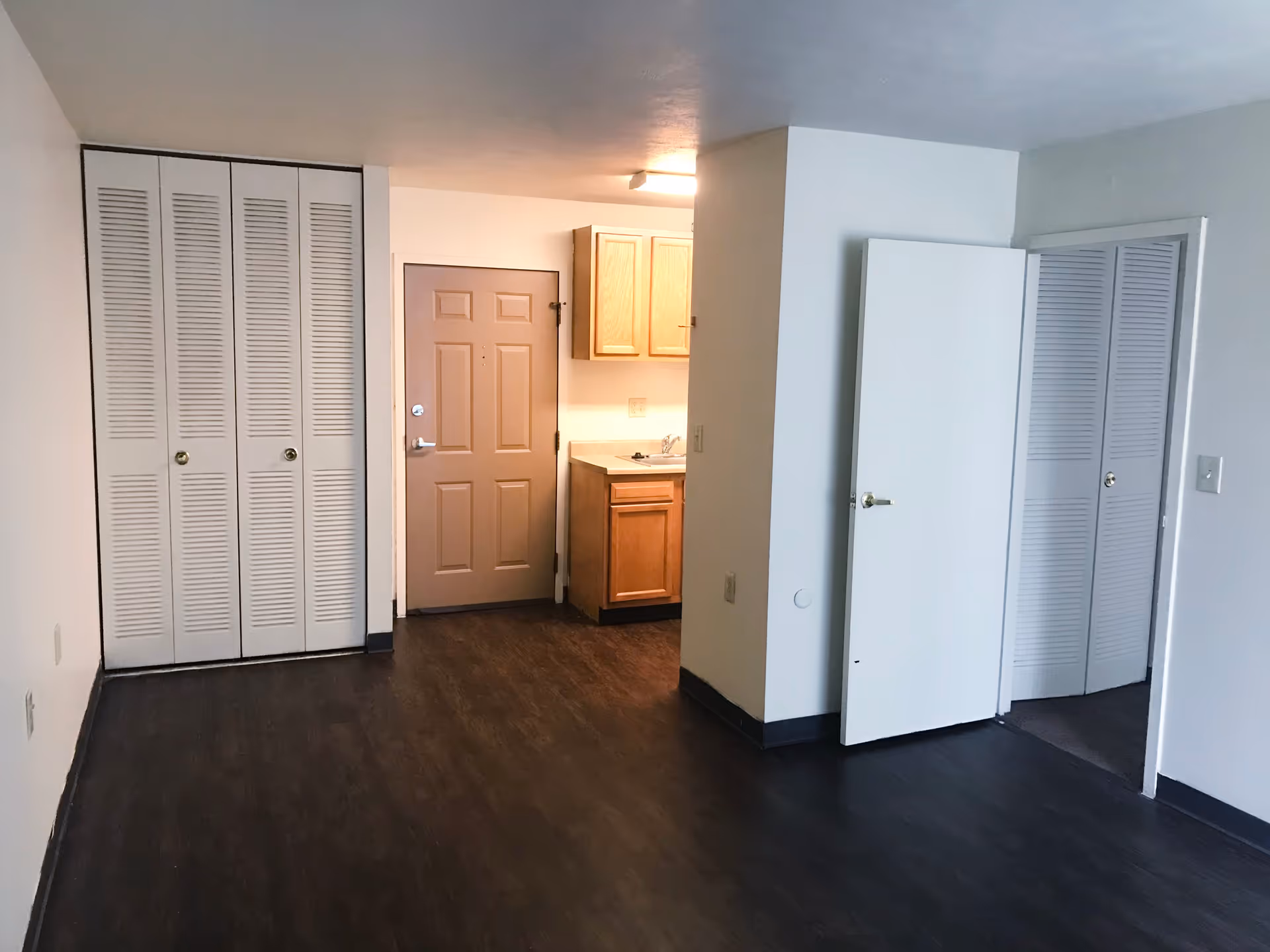 Empty studio apartment interior with an entry door, small kitchenette, closet doors, and dark wood flooring.