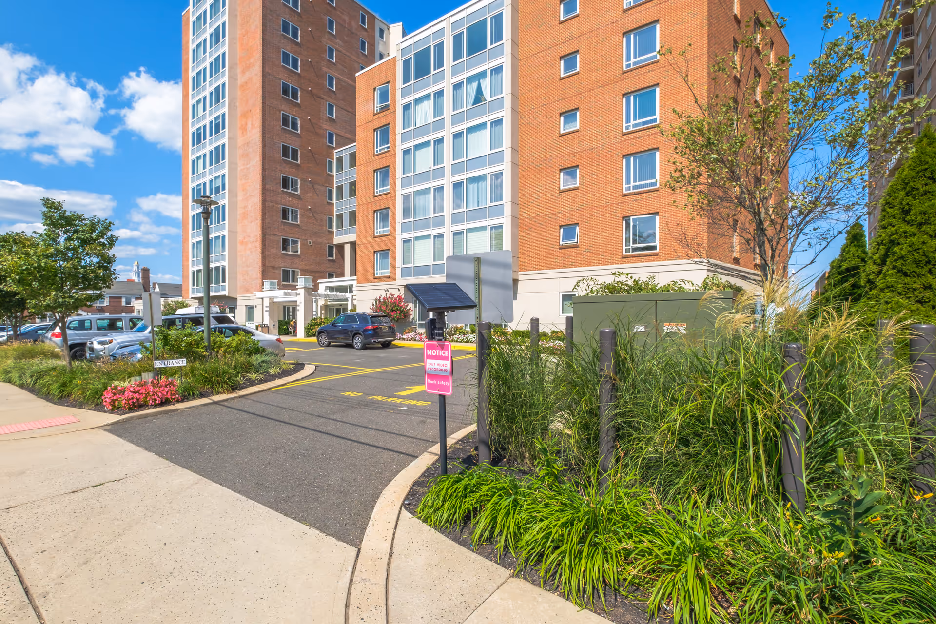 Exterior view of a multi-story brick residential building with large windows, surrounded by landscaped greenery and a parking area with several cars. A sidewalk and a driveway entrance with a sign indicating 24/7 video recording are visible under a bright blue sky with scattered clouds.