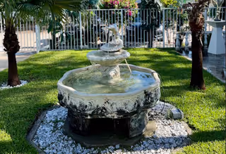 Ornamental tiered stone fountain in a grassy courtyard surrounded by palm trees and a white fence.