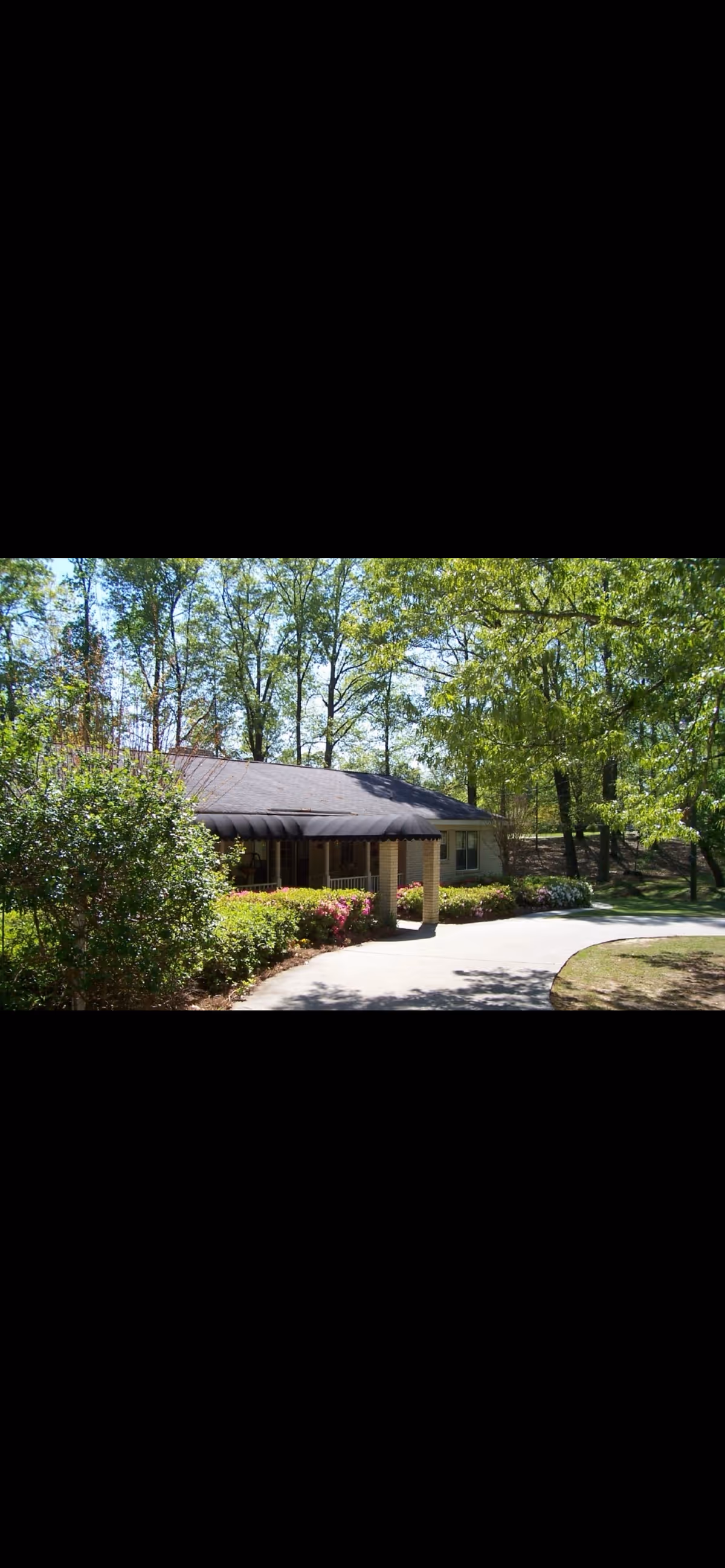 Sunlit entrance of a single-story senior living facility surrounded by trees and shrubs.
