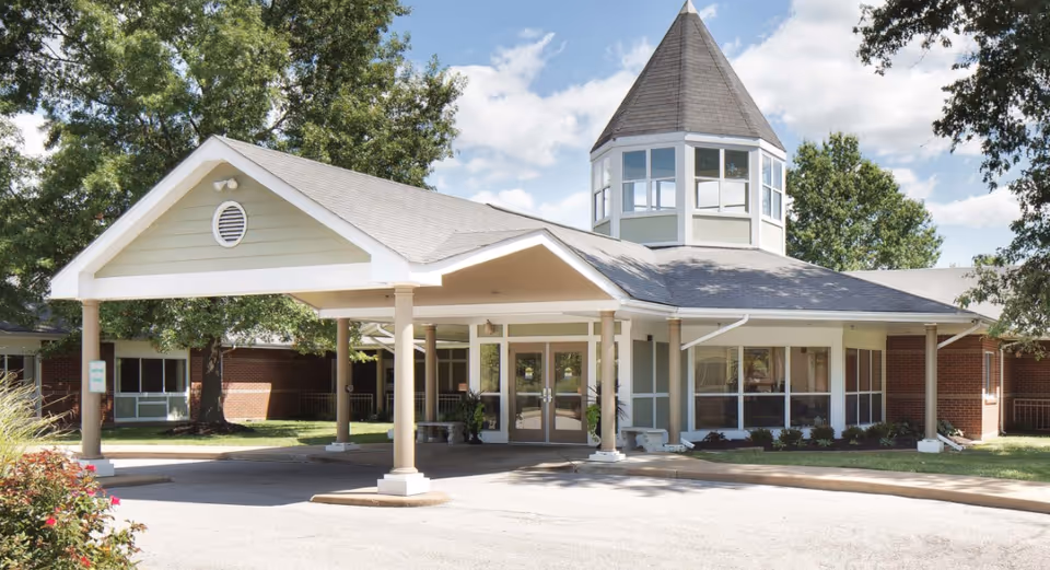 Exterior view of Garden View Care Center of O'Fallon, showing the entrance with a covered driveway supported by columns, a turret-like structure on the roof, and surrounding greenery under a partly cloudy sky.