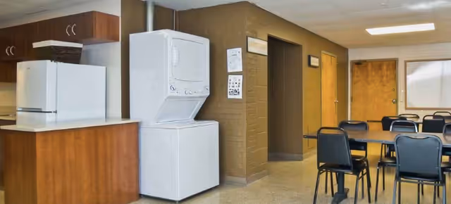 Interior view of a common area in a senior living facility featuring a stacked white washer and dryer next to a wooden cabinet with a white countertop. There are several black chairs and tables arranged in the room, with a door and a bulletin board on the far wall.