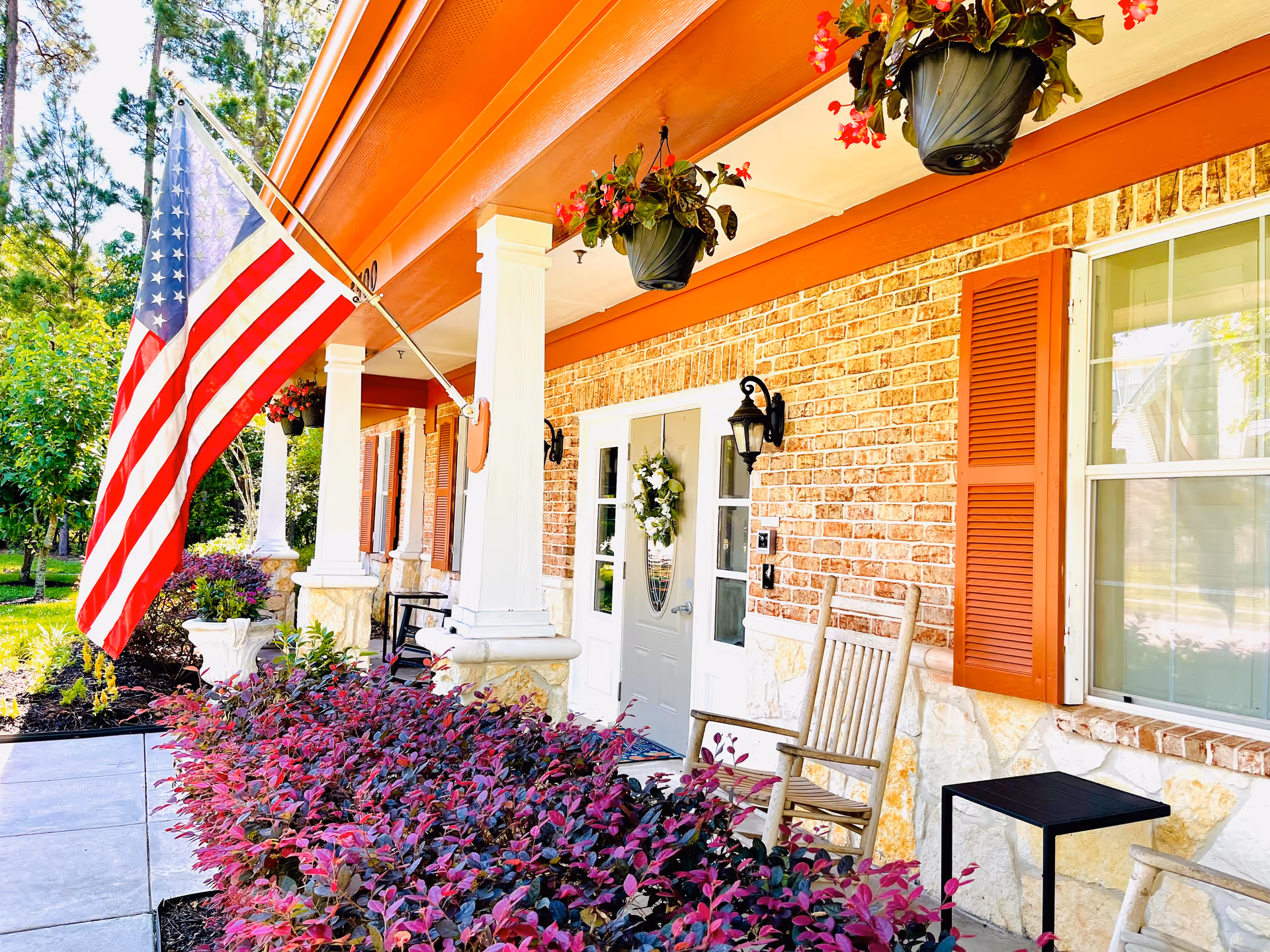 Front porch of a brick cottage showing an American flag, rocking chairs, hanging flower pots, and purple shrubbery.