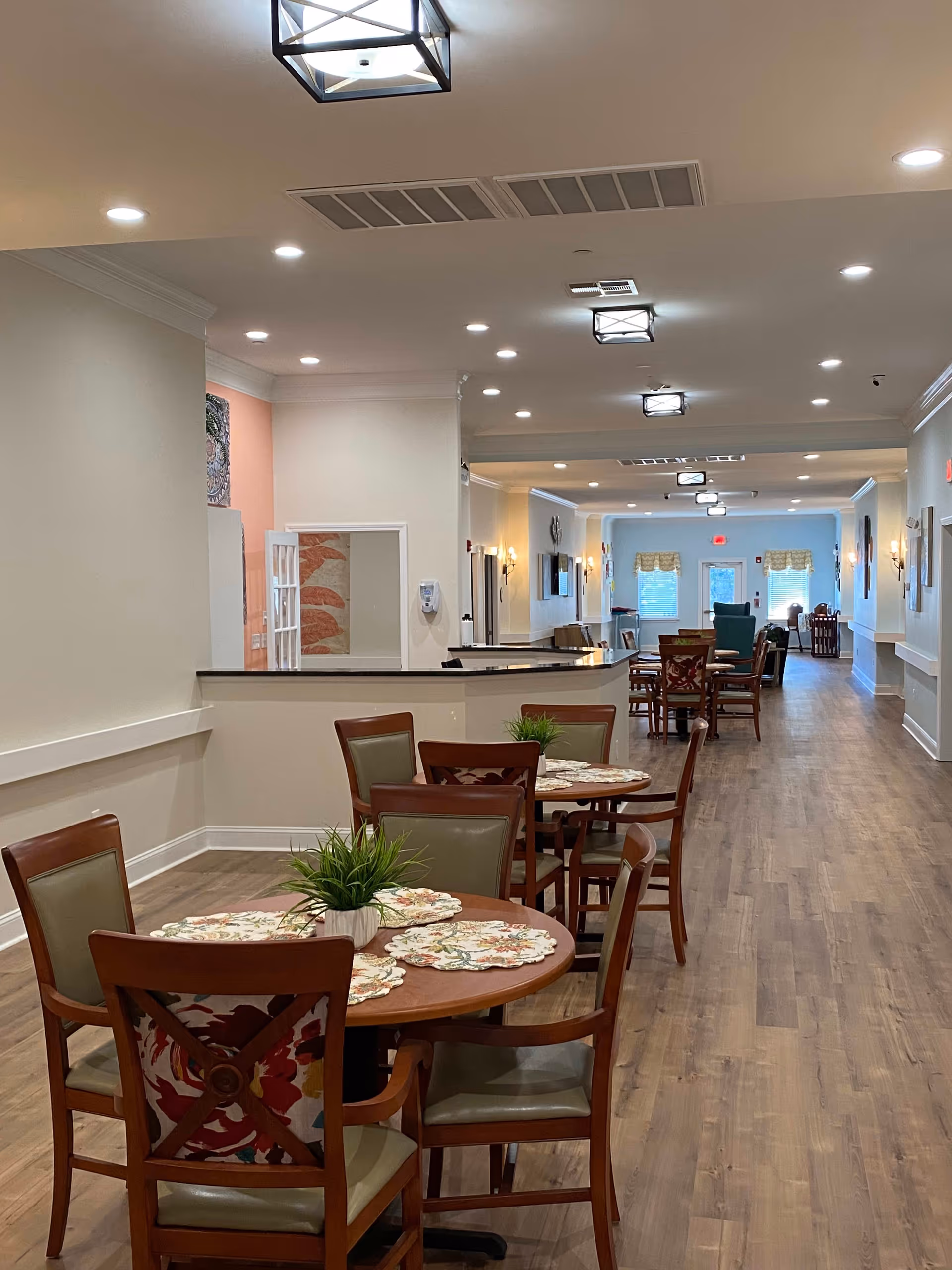 Interior view of a senior living facility dining area with round wooden tables and chairs arranged along a long hallway. The tables have floral placemats and small potted plants. The floor is wood, and the ceiling has recessed lighting and decorative light fixtures. The walls are painted in neutral tones with some artwork and windows at the far end.