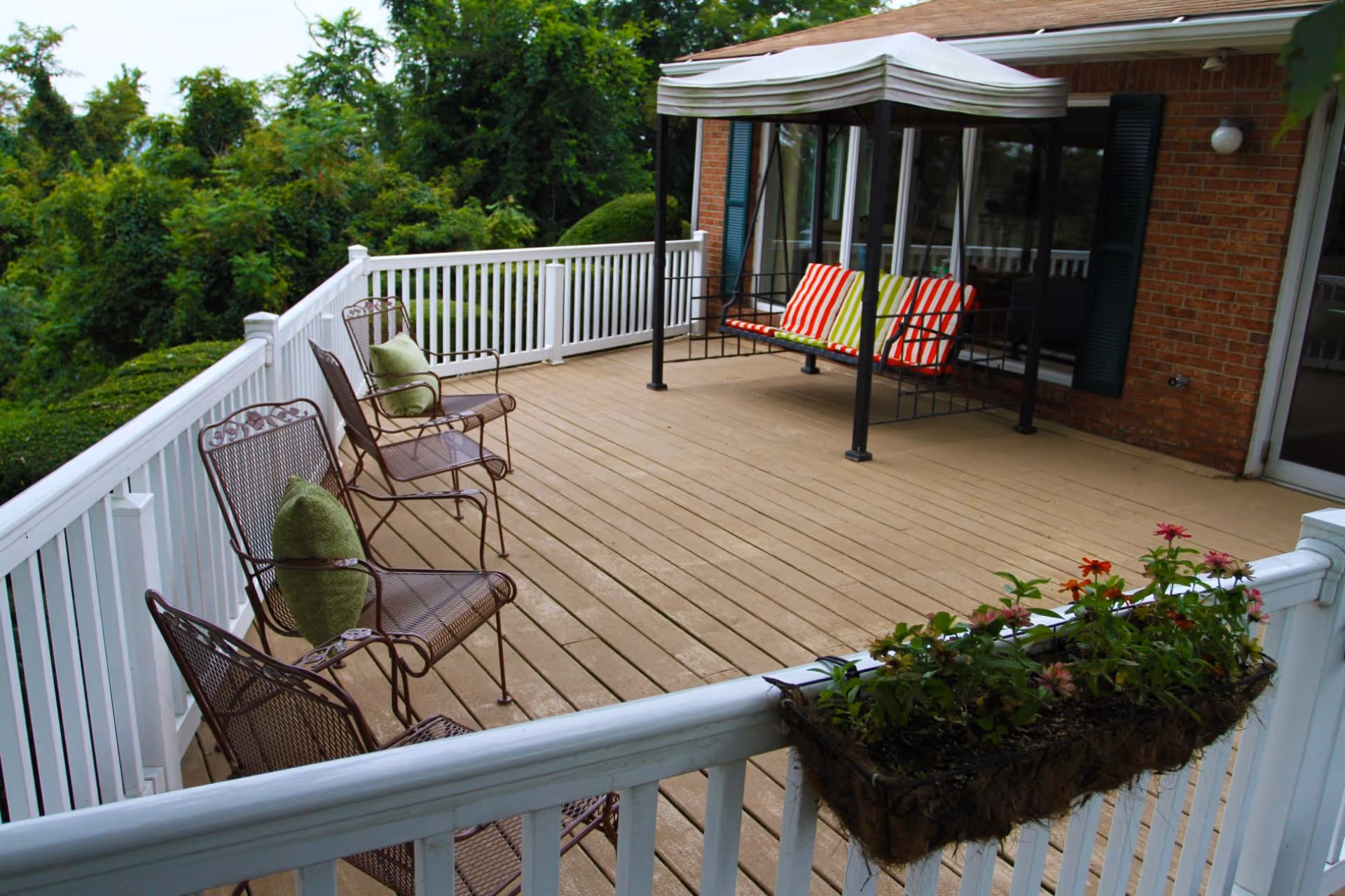 Outdoor patio area with a wooden deck, white railing, three metal chairs with green cushions, a canopy swing with striped cushions, and a flower box with blooming flowers attached to the railing. The patio is adjacent to a brick building with green shutters and surrounded by greenery.