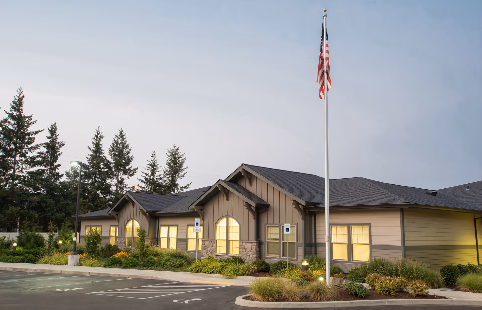 Exterior view of a single-story senior living facility building at dusk with warm lights glowing from the windows. The building has a pitched roof, beige siding with stone accents, and an American flag on a flagpole in front. There are landscaped bushes and plants around the building and a parking lot with handicap parking spaces in the foreground.