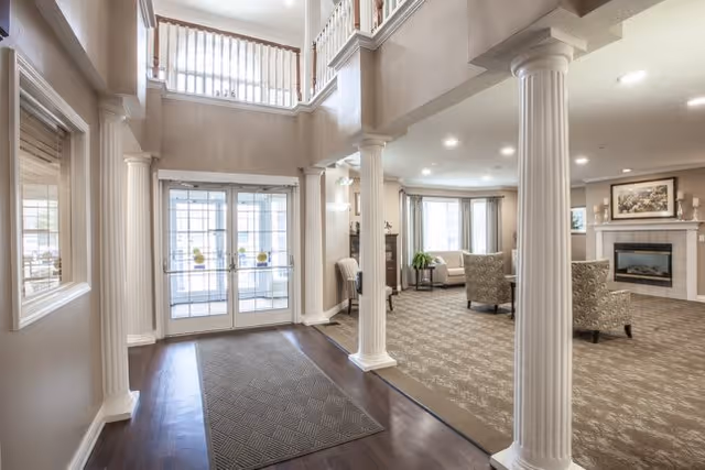 Interior view of a senior living facility lobby area with tall white columns, double glass doors, a seating area with armchairs and a sofa near a fireplace, and a second-floor balcony overlooking the space.