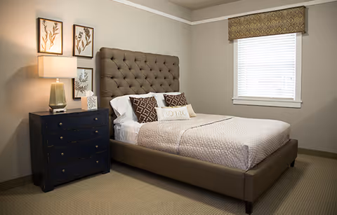 A neatly made bed with a tufted headboard in a bedroom. The bed has white sheets, a beige quilt, and decorative pillows including one with the word 'home'. Next to the bed is a dark blue nightstand with a lamp and a small decorative item. Above the nightstand are three framed botanical prints. A window with a patterned valance is on the right wall.