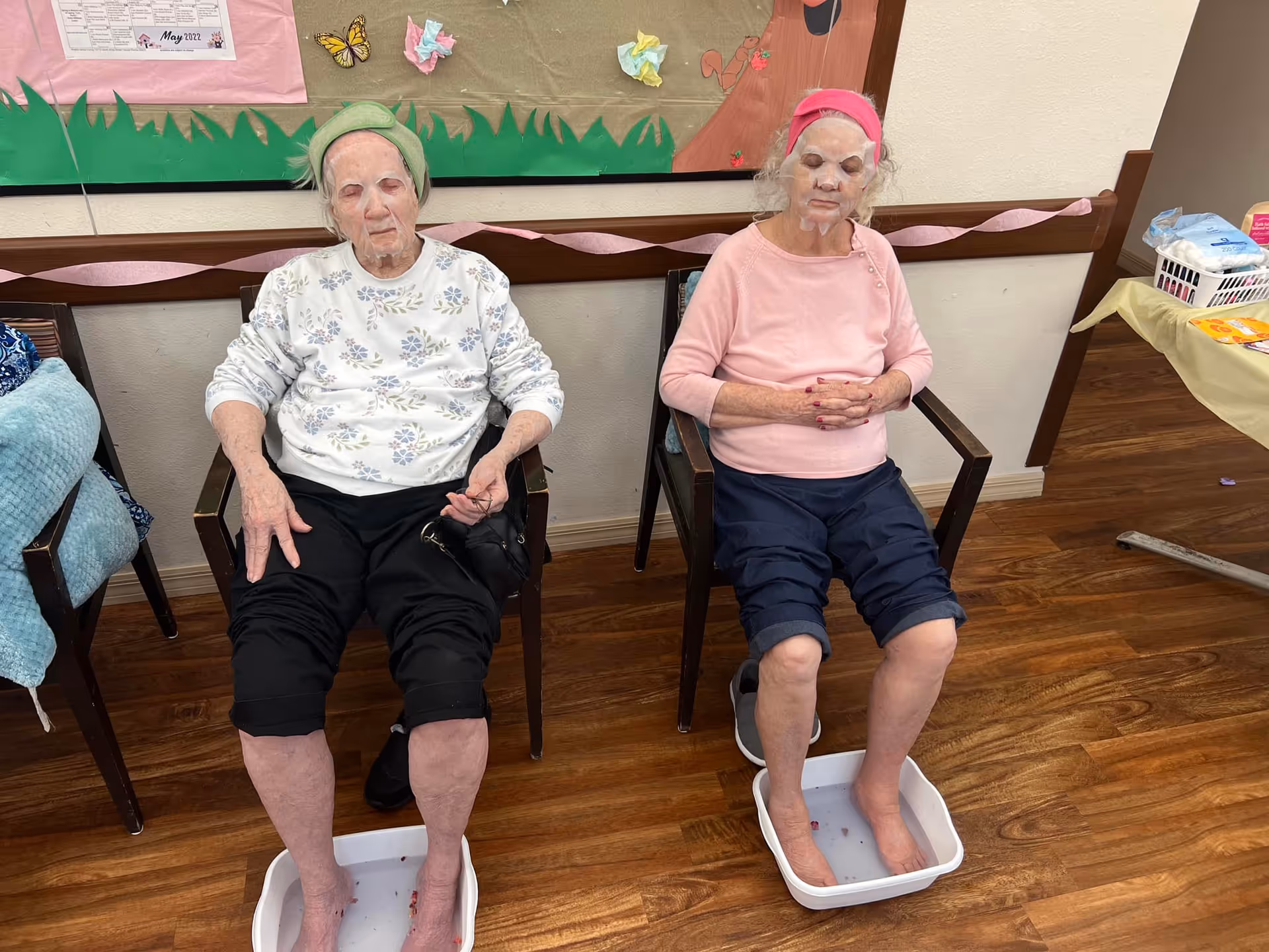 Two elderly women sitting in chairs with their feet soaking in small white tubs filled with water. Both women have facial masks on and are wearing headbands. They appear to be in a communal room with wooden floors and a decorated bulletin board behind them.