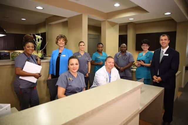A group of healthcare professionals, including nurses and a doctor, standing and sitting behind a reception desk in a medical facility. They are smiling and appear welcoming. The setting is an interior space with warm lighting and a modern design.