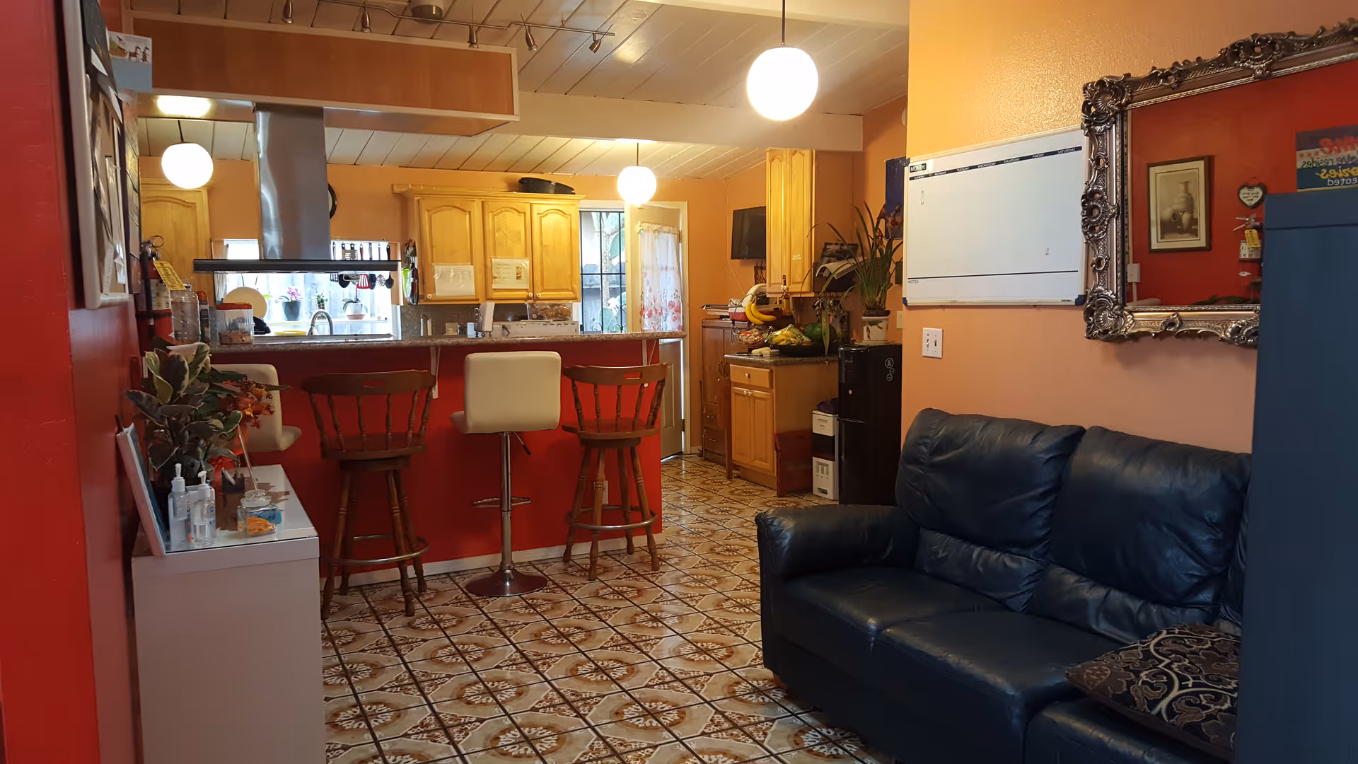 Open living area showing a dark leather couch, a red breakfast bar with three stools, and a kitchen with wooden cabinets in the background.
