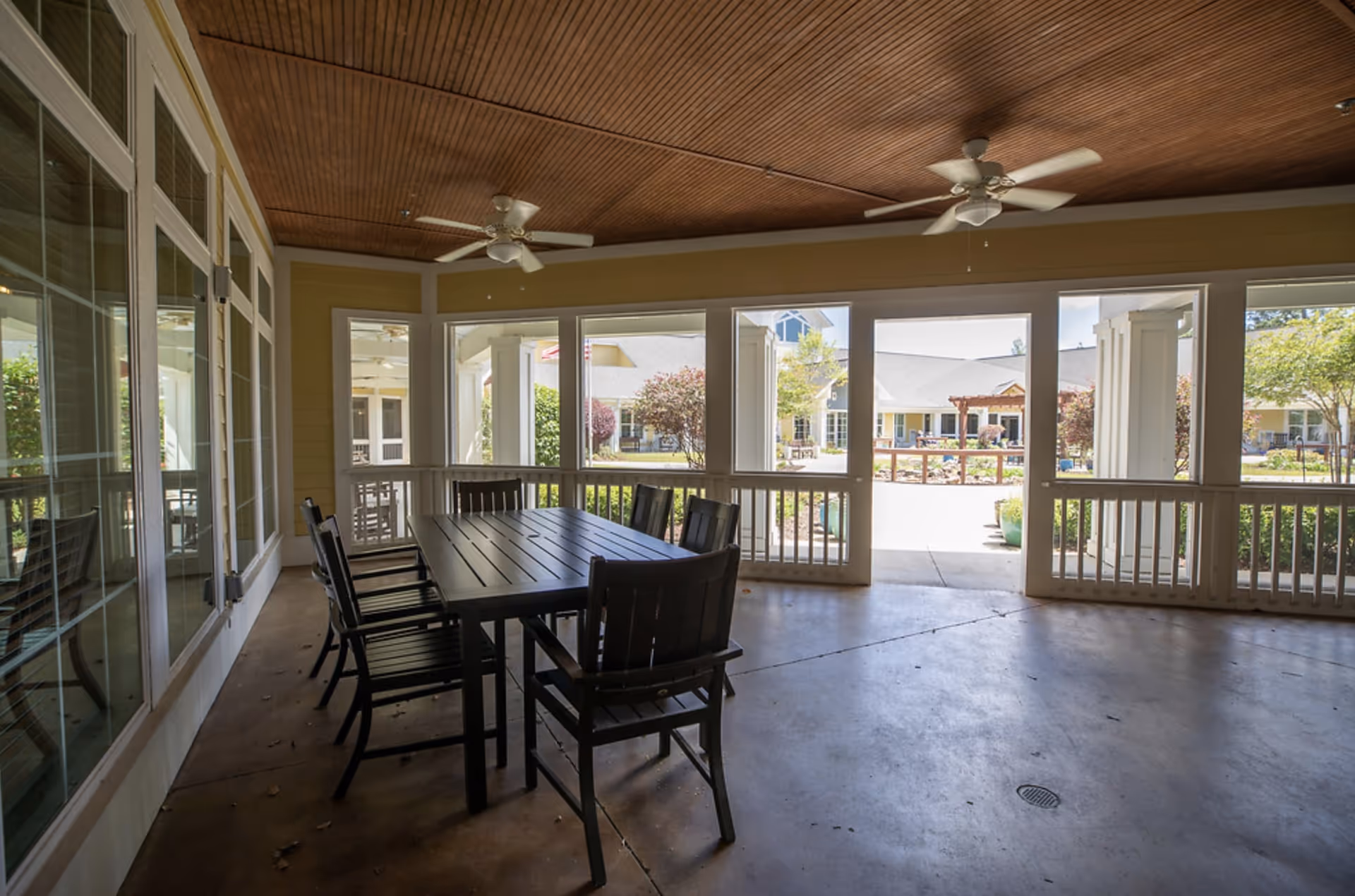 Covered outdoor patio area with a wooden ceiling, two ceiling fans, and a black dining table surrounded by six black chairs. The patio overlooks a courtyard with greenery and buildings in the background.