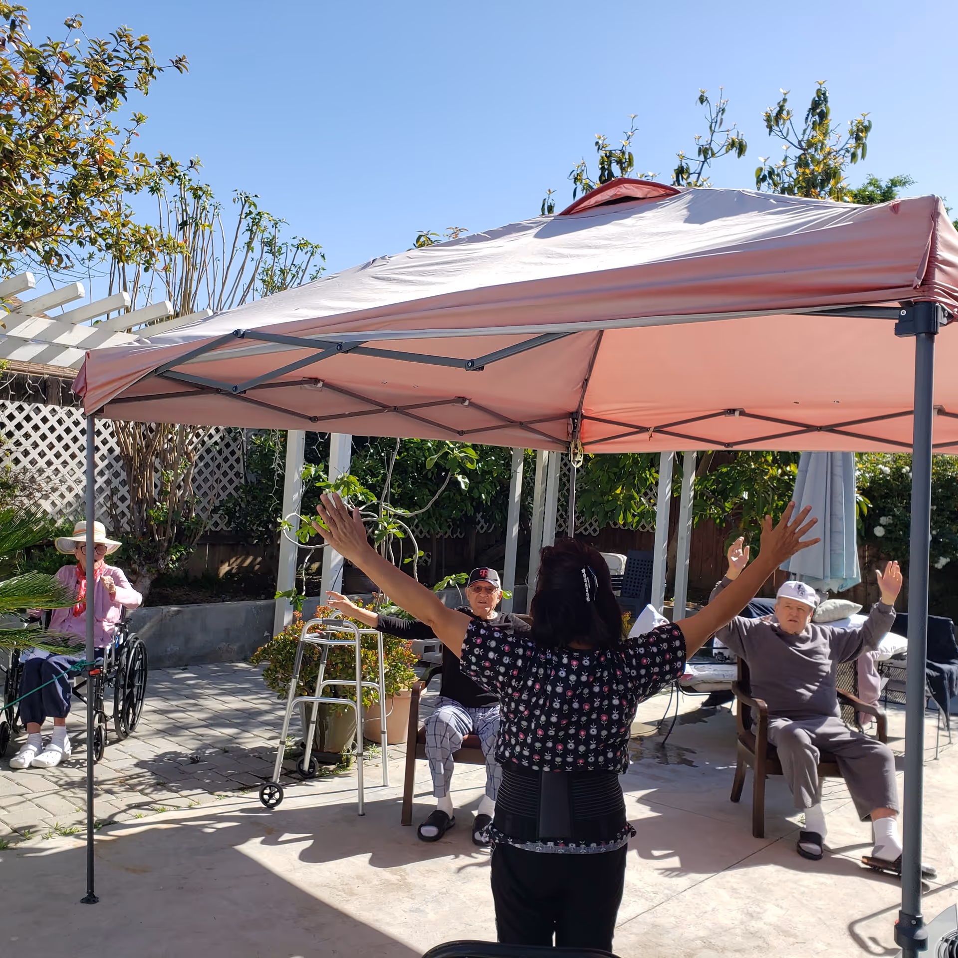 An outdoor patio area at Terrace Gardens with a pink canopy providing shade. Three elderly individuals are seated in chairs, participating in a group activity led by a woman standing with her back to the camera, raising her arms. The setting is sunny with clear blue skies and greenery surrounding the patio.