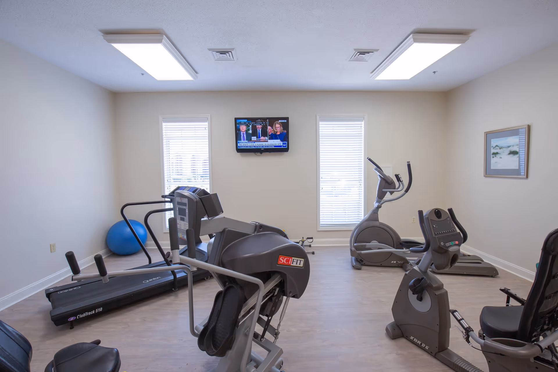 A small fitness room with exercise equipment including a treadmill, elliptical machine, stationary bike, and a strength training machine. The room has light-colored walls, two windows with blinds, a wall-mounted TV, and a framed picture on the wall.