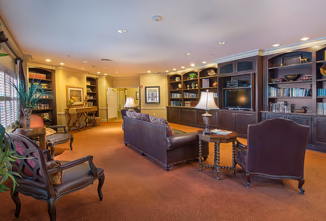 A warm, carpeted communal lounge with leather sofas and chairs, built-in bookcases and a TV, lit by table lamps and recessed ceiling lights.