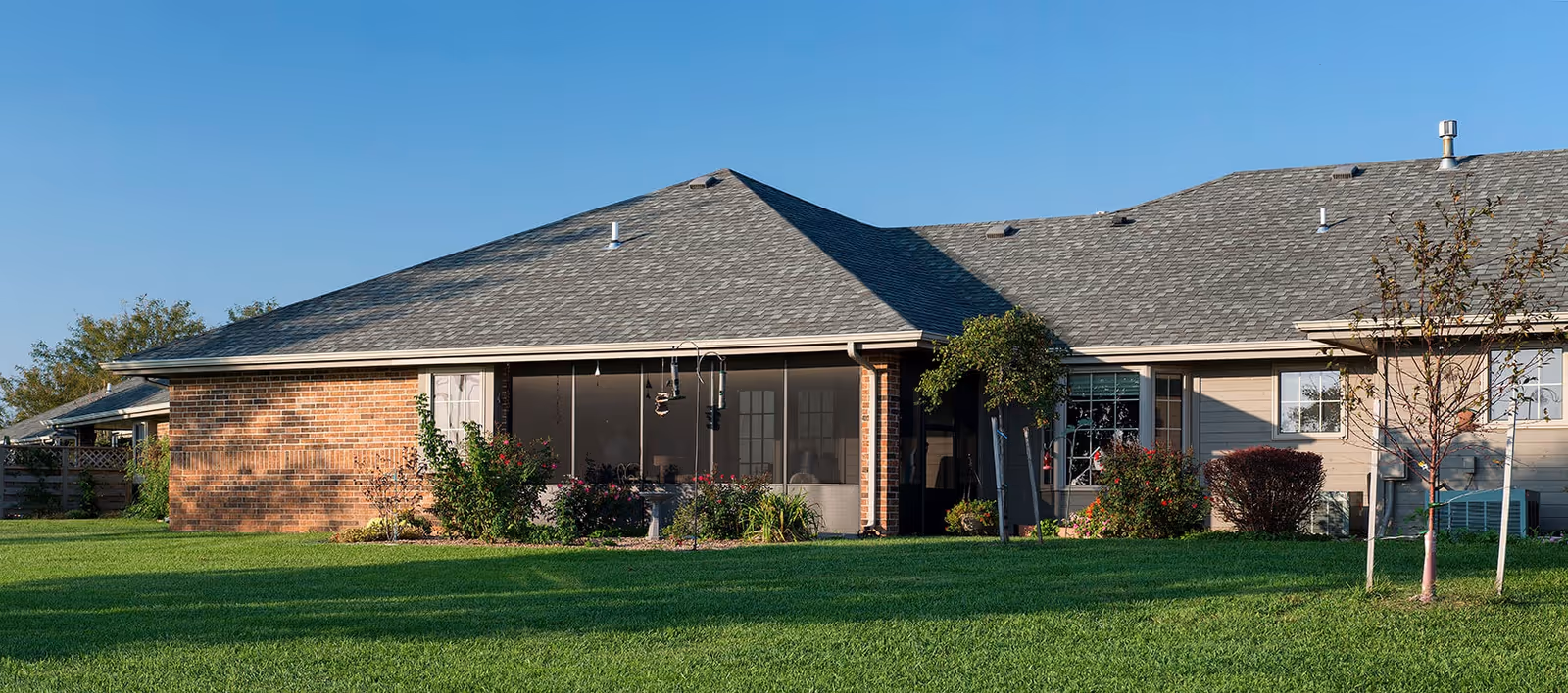 Exterior view of a single-story residential building with a gray shingled roof, brick and siding walls, a screened porch, and a well-maintained green lawn with some shrubs and small trees.