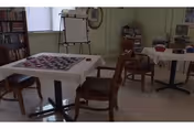 A room with several tables and chairs arranged for activities. One table has a checkerboard set up for a game. There is a bookshelf filled with books, a whiteboard on a stand, and various other items suggesting a communal or activity space.