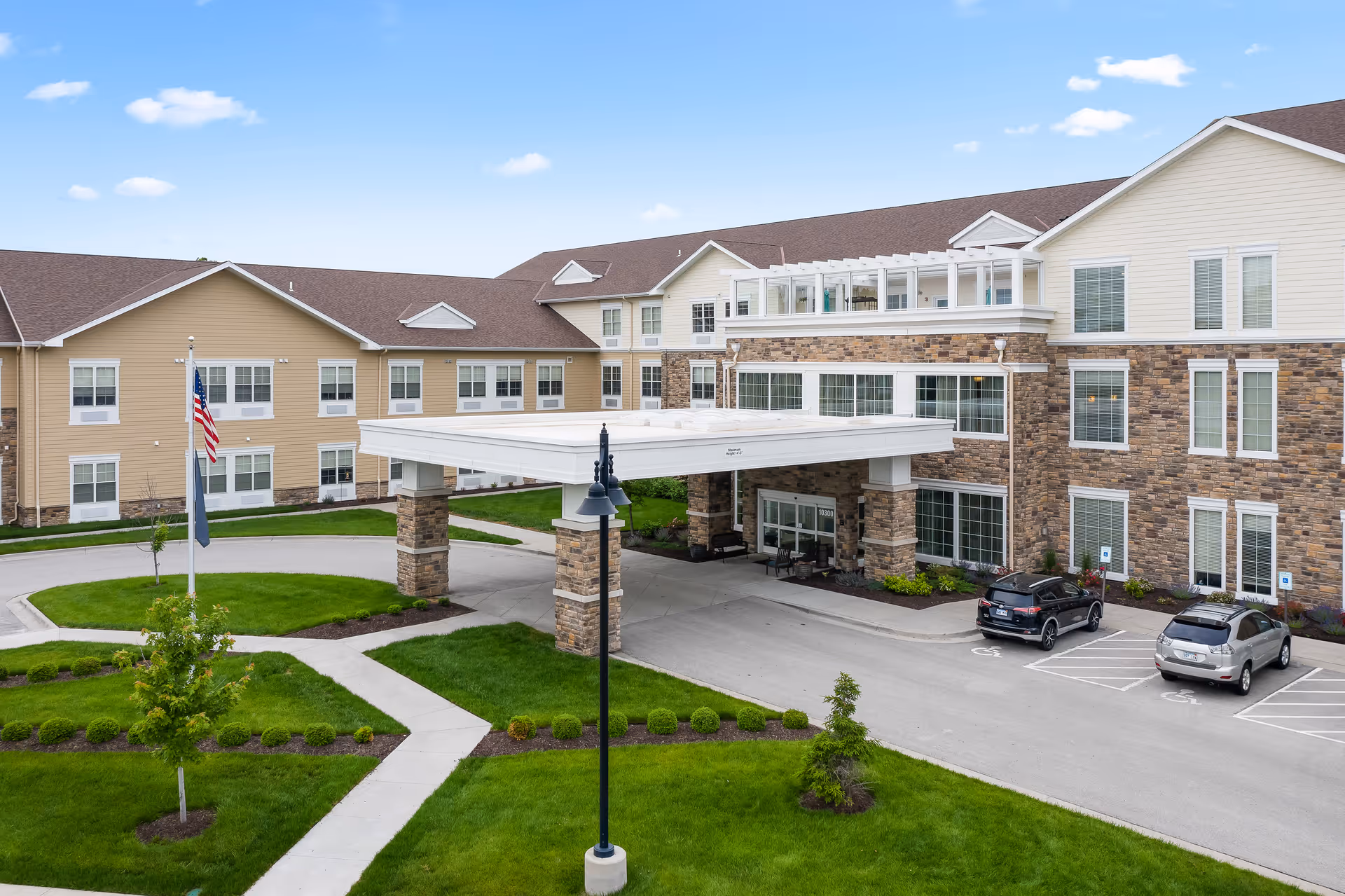 Exterior view of a senior living facility named Alto Overland Park featuring a large covered entrance supported by stone pillars, a well-maintained lawn with pathways, an American flag, and a parking area with two cars.