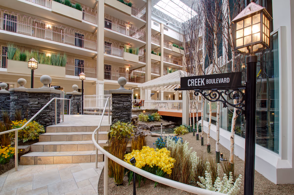 Indoor courtyard area of a senior living facility featuring a tiled staircase with metal railings, stone pillars, decorative street lamps, and a garden with yellow and white flowers. A black sign reads 'CREEK BOULEVARD'. The background shows multiple floors with balconies and plants.