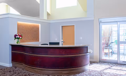Reception area with a curved wooden desk featuring decorative nailhead trim, a vase of flowers on the counter, a closed wooden door behind the desk, and glass sliding doors to the right leading outside.