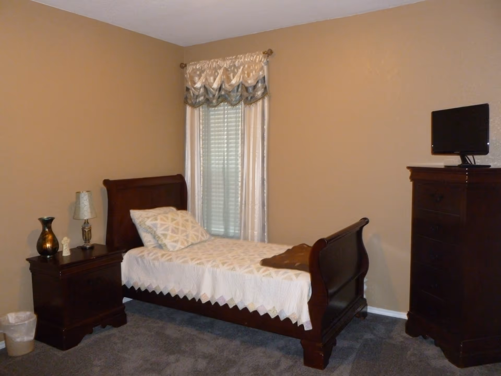 A bedroom with a single wooden bed covered with a white quilt and a patterned pillow. Next to the bed is a wooden nightstand with a lamp, a decorative vase, and a small figurine. A window with white and blue curtains is behind the bed. On the right side of the room, there is a tall wooden dresser with a small flat-screen TV on top. The walls are beige and the floor is carpeted in gray.