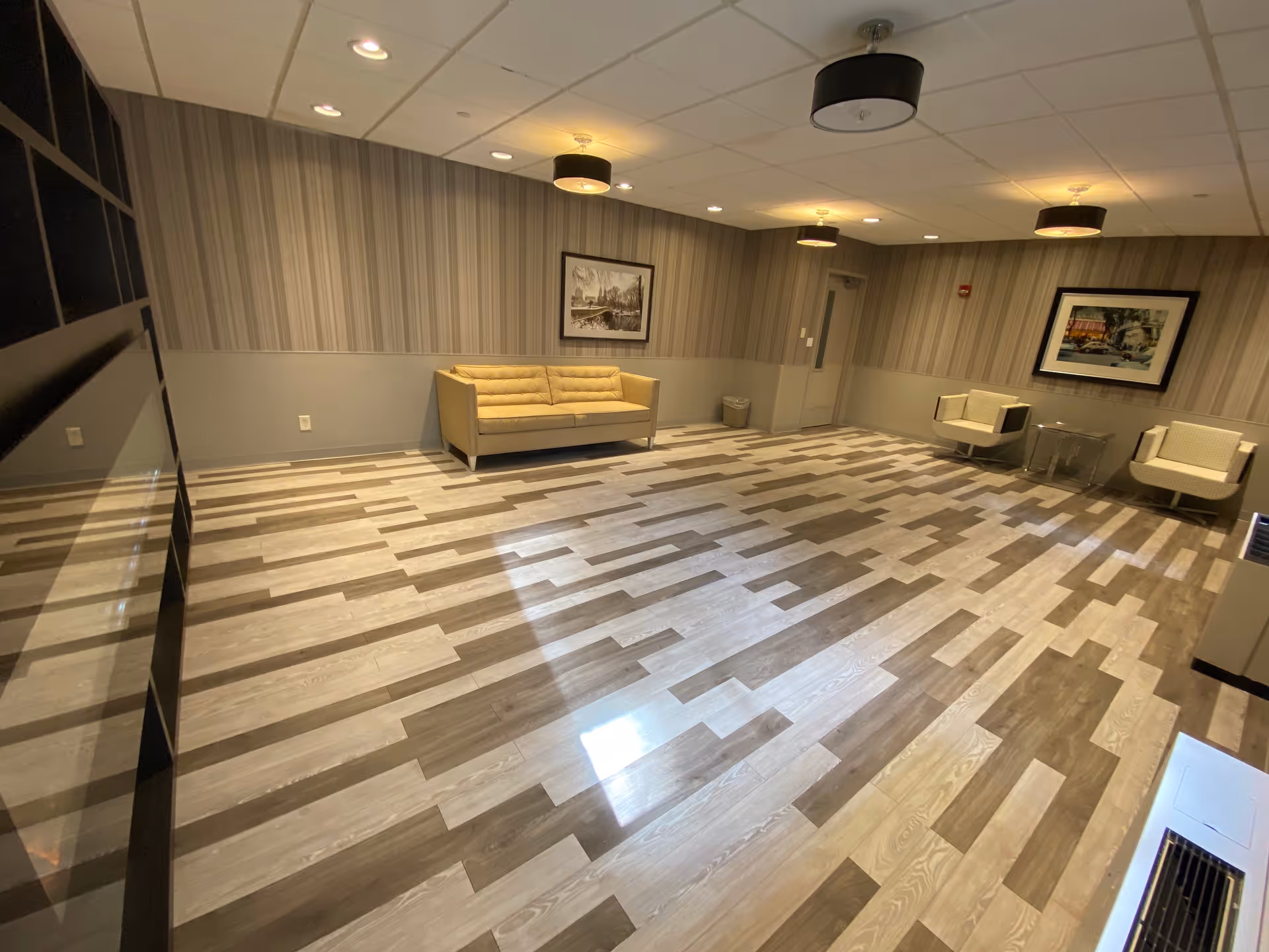 A spacious, modern lounge area with patterned wood flooring, beige sofa, two beige armchairs, two framed pictures on the walls, and ceiling lights. The walls have a two-tone design with striped wallpaper on the upper half and solid color on the lower half.