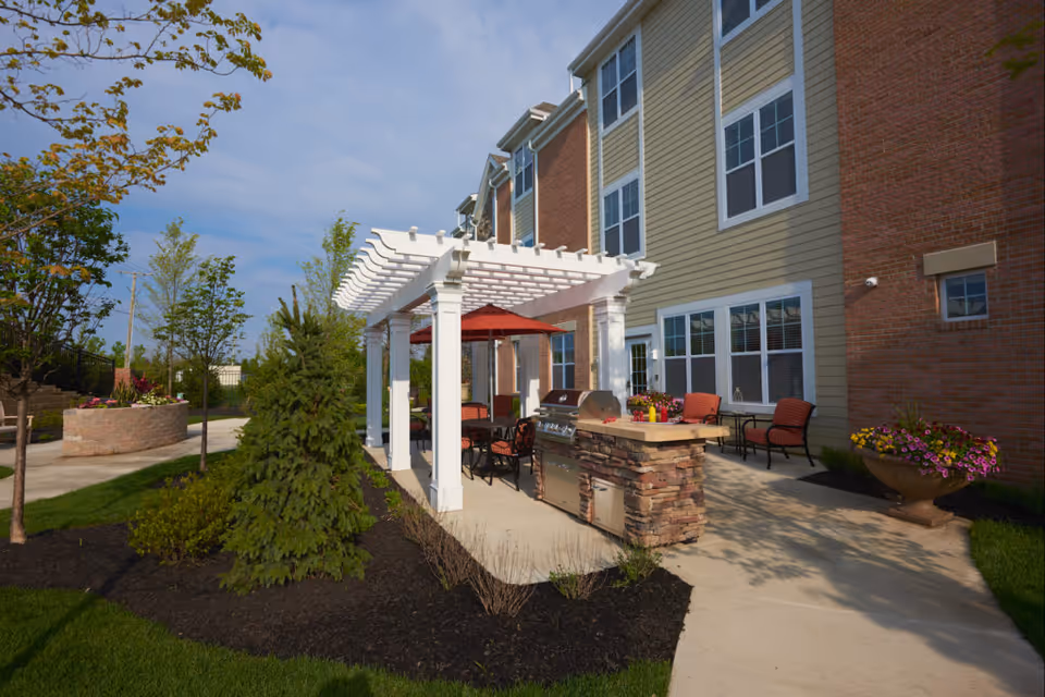 Outdoor patio area with a white pergola, built-in grill, seating and planters alongside a multi-story retirement building.