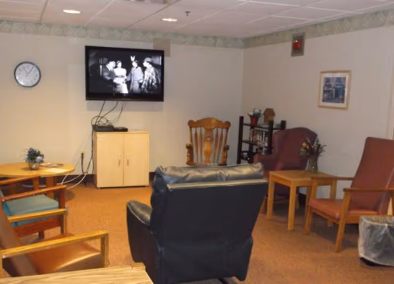 Small communal lounge with several chairs and tables facing a wall-mounted TV showing a black-and-white film.