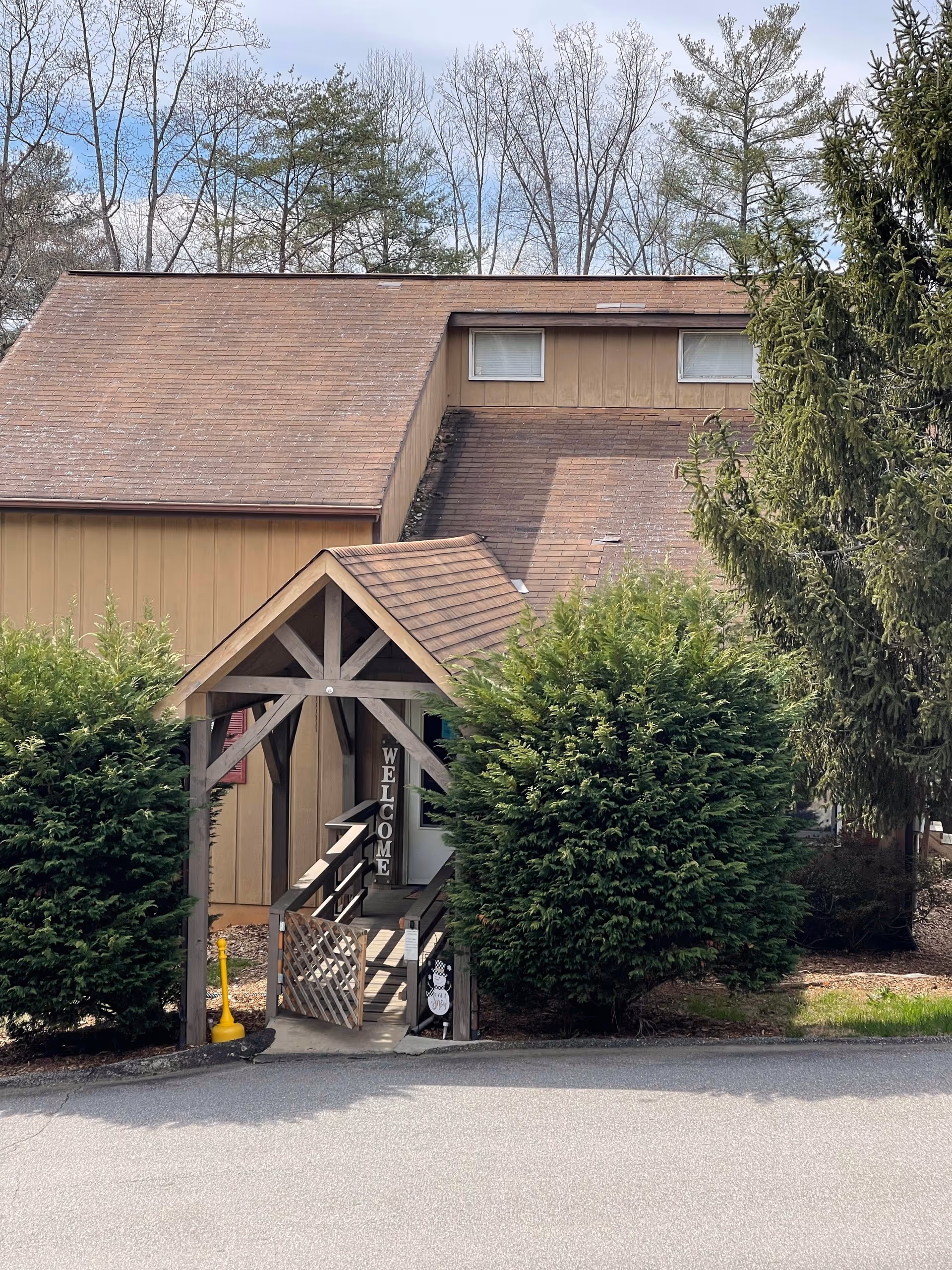 Exterior view of a building with a brown sloped roof and beige siding, partially obscured by green bushes and trees. A wooden covered entrance with a ramp and a gate is visible, with a vertical 'WELCOME' sign hanging inside the entrance.