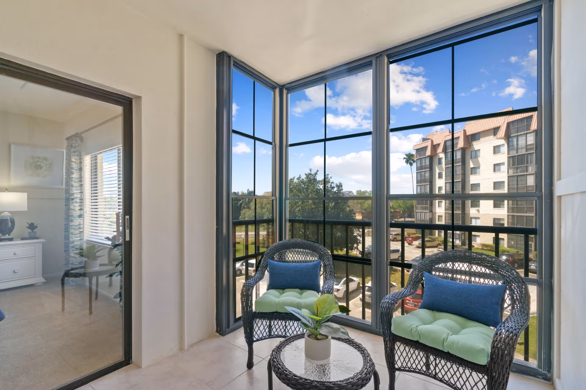 Sunlit enclosed balcony/sunroom with two wicker chairs, a small table, and large windows overlooking a parking lot and neighboring building.