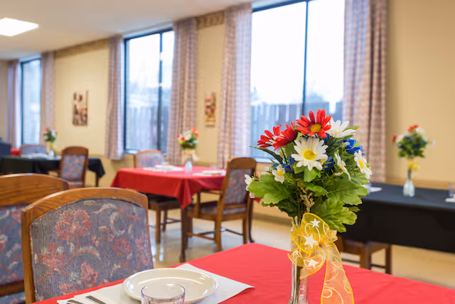 Dining room with tables covered in red and black tablecloths, each table decorated with a vase of colorful flowers. Chairs with floral upholstery surround the tables, and large windows with patterned curtains let in natural light.