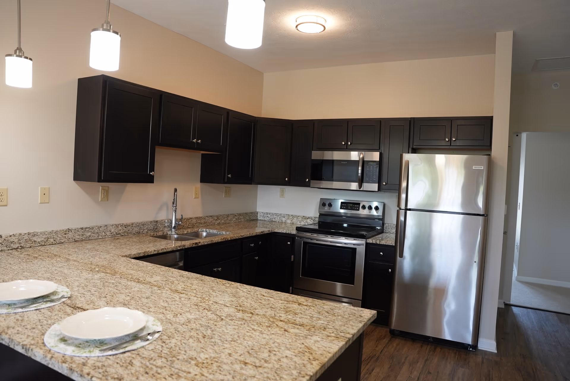 Modern kitchen with dark wood cabinets, granite countertops, stainless steel appliances including a refrigerator, oven, and microwave. Two pendant lights hang above the counter, which has two place settings with plates and placemats.