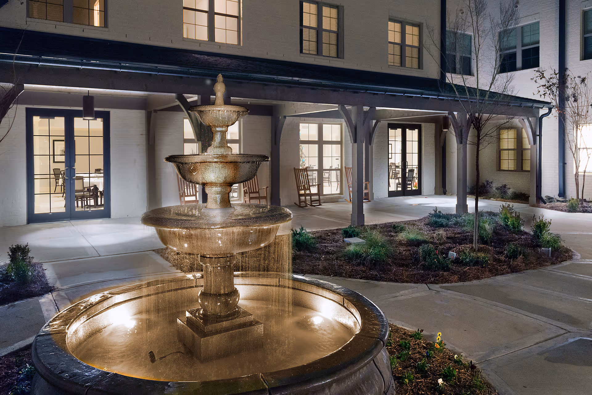 Night view of an outdoor courtyard at The Magnolia at Oxford Commons featuring a three-tiered water fountain in the foreground, surrounded by landscaped plants and a concrete walkway. The building in the background has large windows and doors with rocking chairs on the covered porch area.