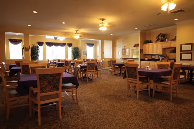 A spacious dining room with multiple wooden tables and chairs, each table covered with a dark tablecloth. The room is warmly lit with ceiling lights and ceiling fans. There are windows with valances letting in natural light, and some greenery and framed pictures decorate the walls. A kitchenette area with cabinets and a counter is visible on the right side.