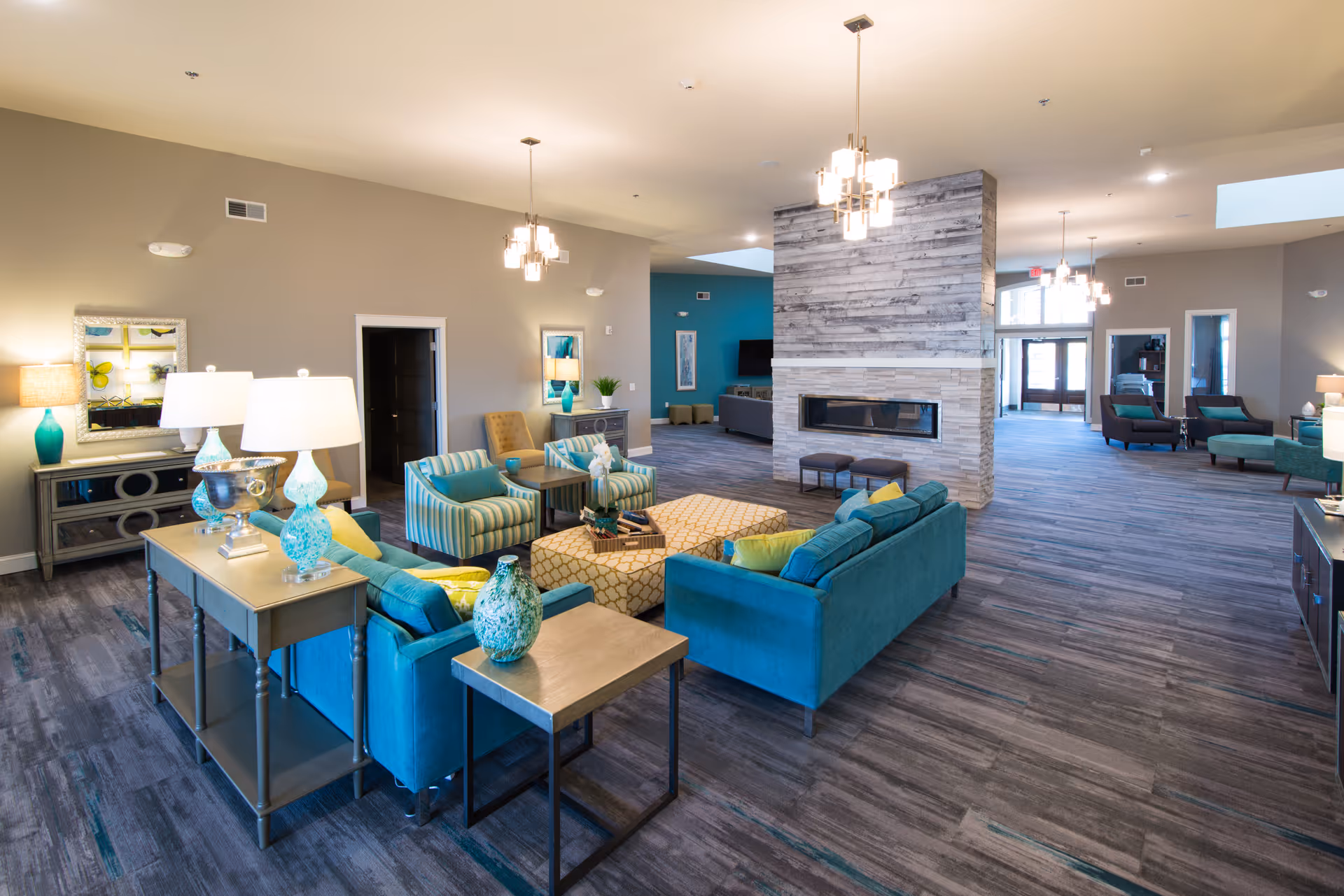 A spacious and modern living room area in a senior living facility with teal and yellow upholstered sofas and chairs arranged around a central ottoman. The room features a large stone fireplace in the center, multiple pendant lights hanging from the ceiling, and a mix of wooden and glass furniture. The flooring is a dark wood pattern, and the walls are painted in neutral tones with some teal accent walls. There are lamps and decorative vases on tables, and the room has a bright and welcoming atmosphere.