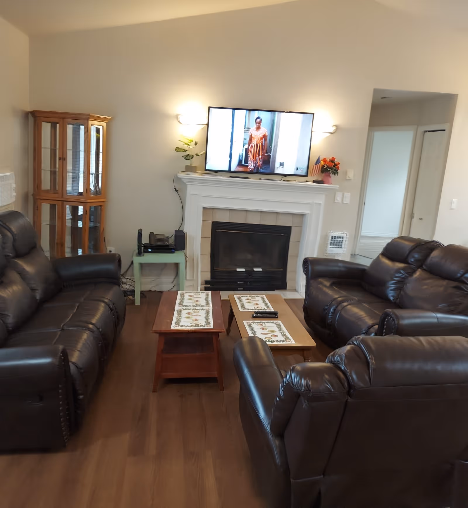 A living room with three dark brown leather recliner sofas arranged around two wooden coffee tables with decorative runners. A white fireplace with a mounted flat-screen TV above it is visible, showing a person in an orange dress. A small green side table with electronic devices and a wooden glass-front cabinet are also present. The room has wooden flooring and beige walls.
