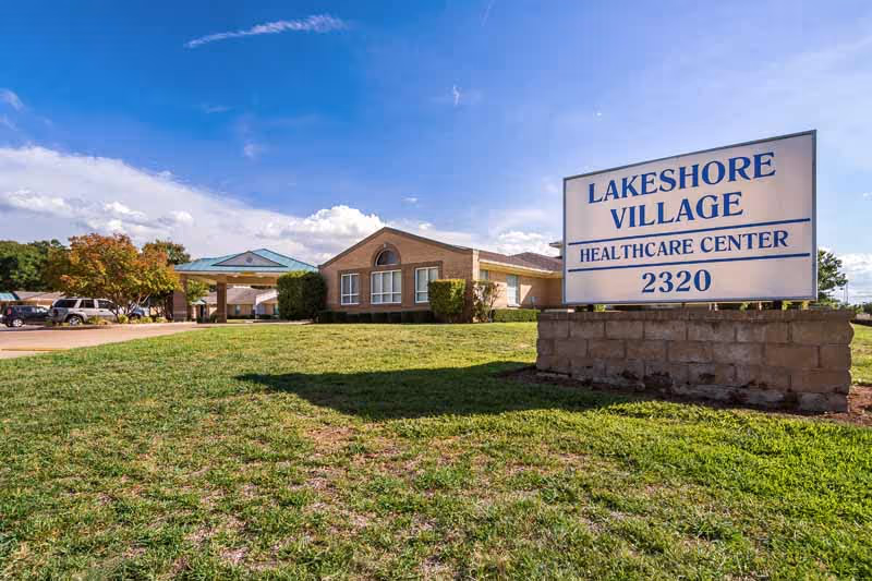 Exterior view of Lakeshore Village Healthcare Center building with a large sign displaying the facility name and address 2320, surrounded by green grass and a clear blue sky.