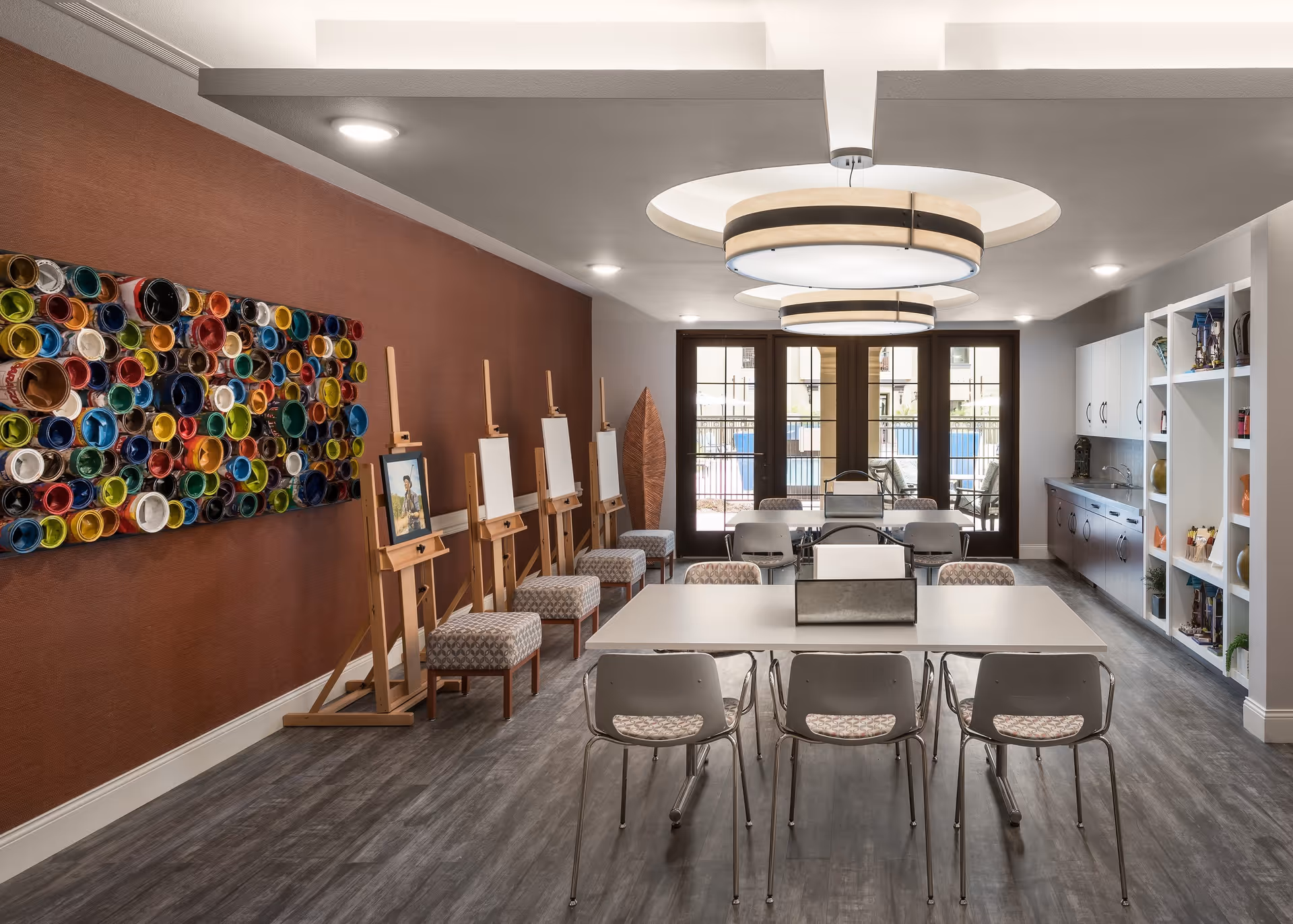 A bright and modern art room with several easels and chairs arranged along a reddish-brown wall. A colorful circular wall art installation is displayed on the left wall. The room features two large circular ceiling lights, a long table with chairs in the center, and a set of glass doors at the far end leading outside. On the right side, there are white cabinets and shelves with decorative items.