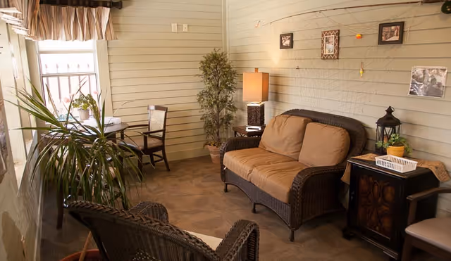 Cozy sunroom-style sitting area with a wicker loveseat and chairs, side tables, lamp, and potted plants.