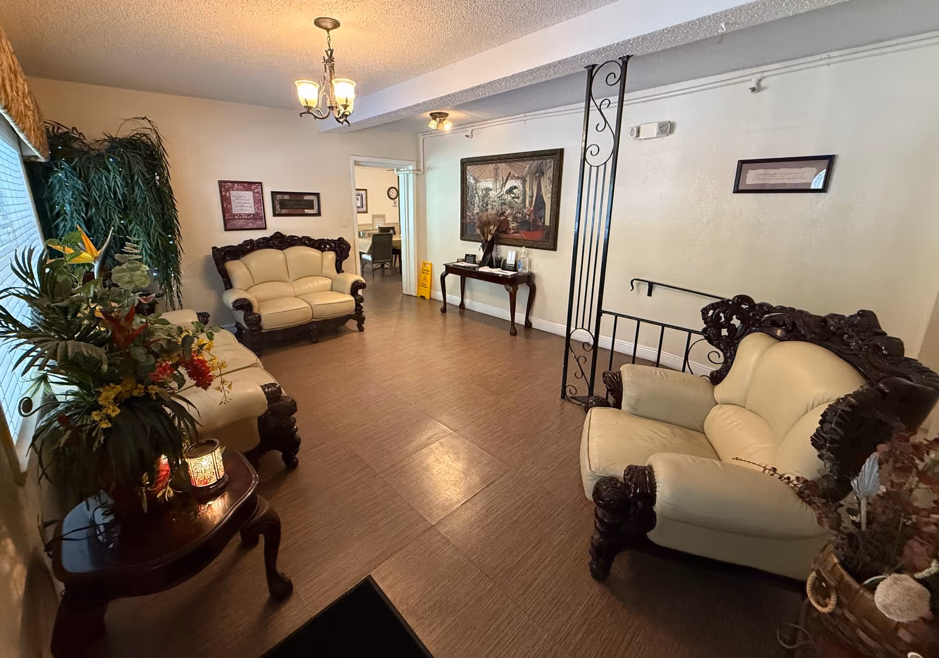 A cozy living room area with cream-colored leather sofas featuring dark wooden frames. There are decorative plants and flowers on a small wooden table near a window with blinds. The room has wooden flooring, a chandelier light fixture, framed artwork on the walls, and a small table with decorative items against one wall.
