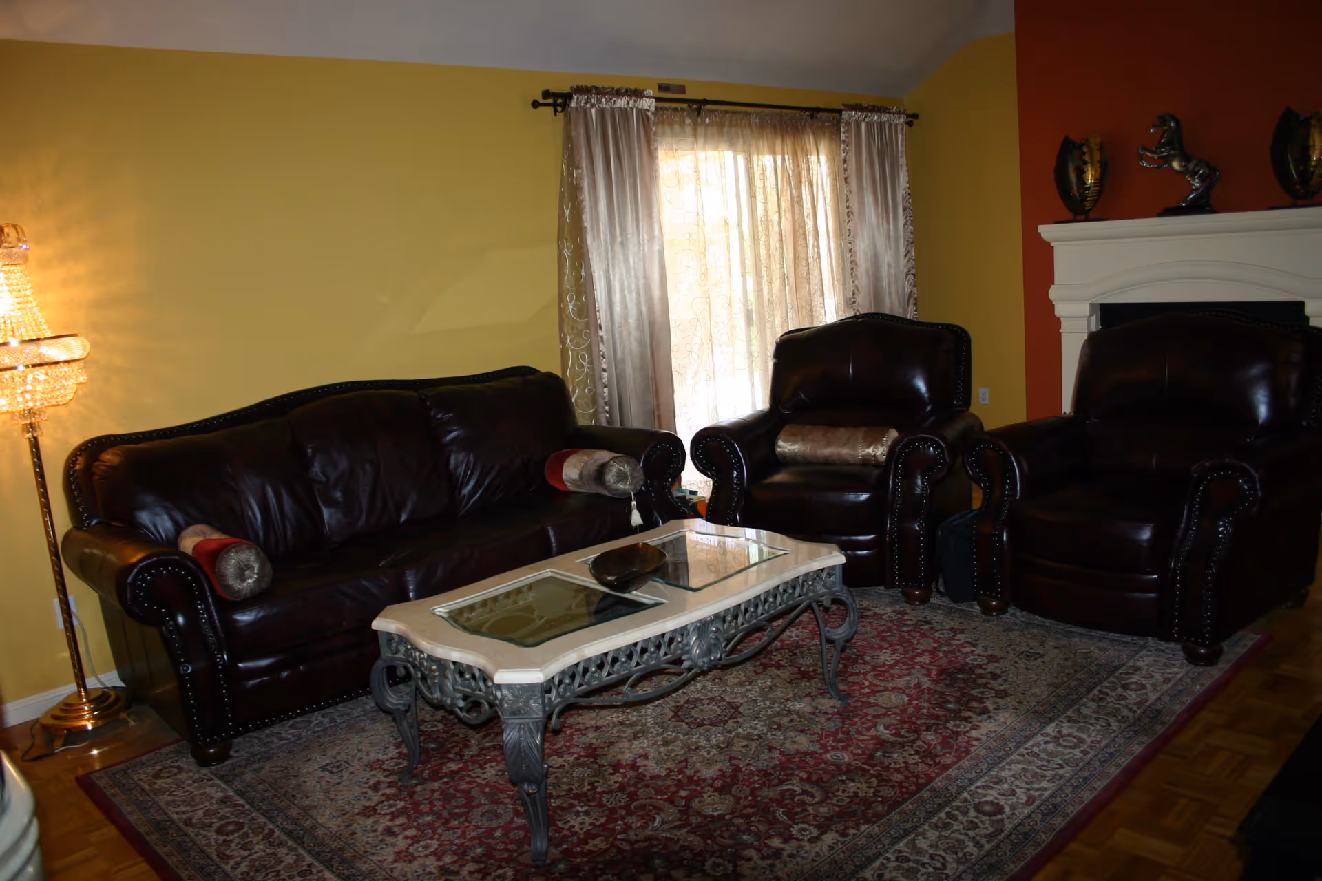 A cozy living room with dark brown leather sofa and two matching armchairs arranged around a decorative glass-top coffee table on a patterned area rug. The walls are painted yellow and orange, with a window covered by sheer curtains letting in natural light. A crystal floor lamp stands to the left, and a white fireplace mantel with decorative items is on the right.