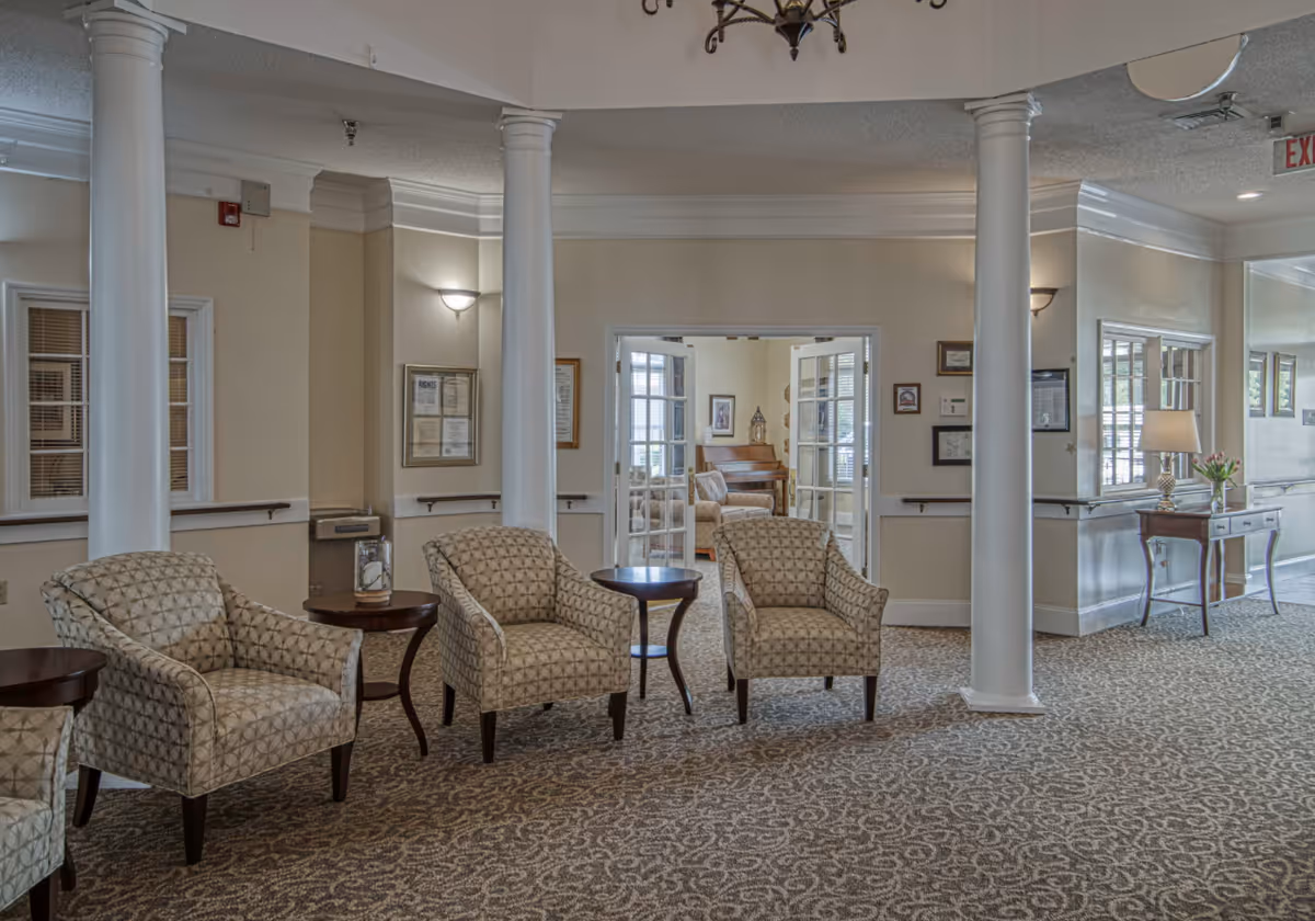 A seating area in a senior living facility with patterned armchairs arranged around small wooden tables. The space features white columns, beige walls, and carpeted flooring with a decorative pattern. In the background, there is a room with French doors, a piano, and additional seating. The area is well-lit with wall sconces and a chandelier.