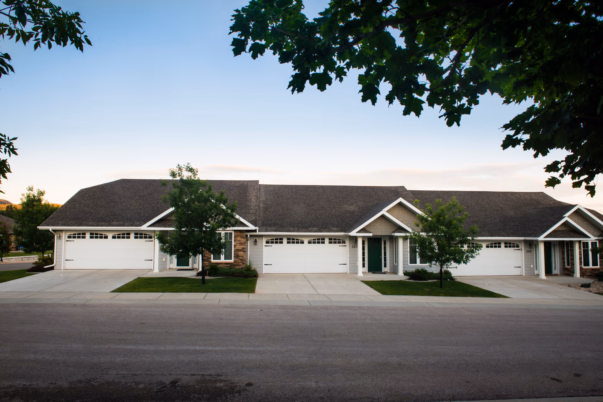 Exterior view of a single-story senior living facility building with multiple attached units, each featuring a garage door and a front entrance. The building has a sloped roof and is surrounded by small trees and a paved driveway. The sky is clear with a hint of sunset light.