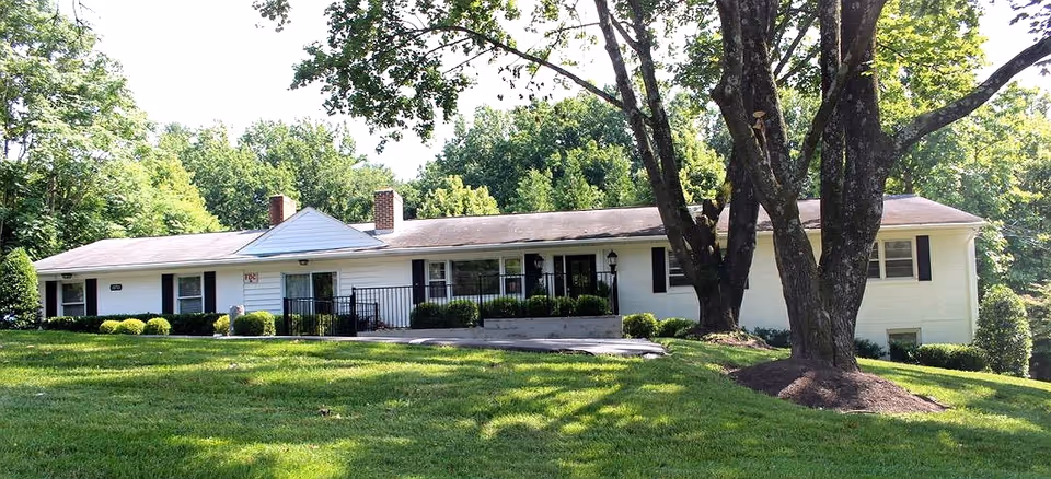 Single-story white building with black shutters surrounded by green trees and a well-maintained lawn, with a large tree in the foreground casting shadows on the grass.