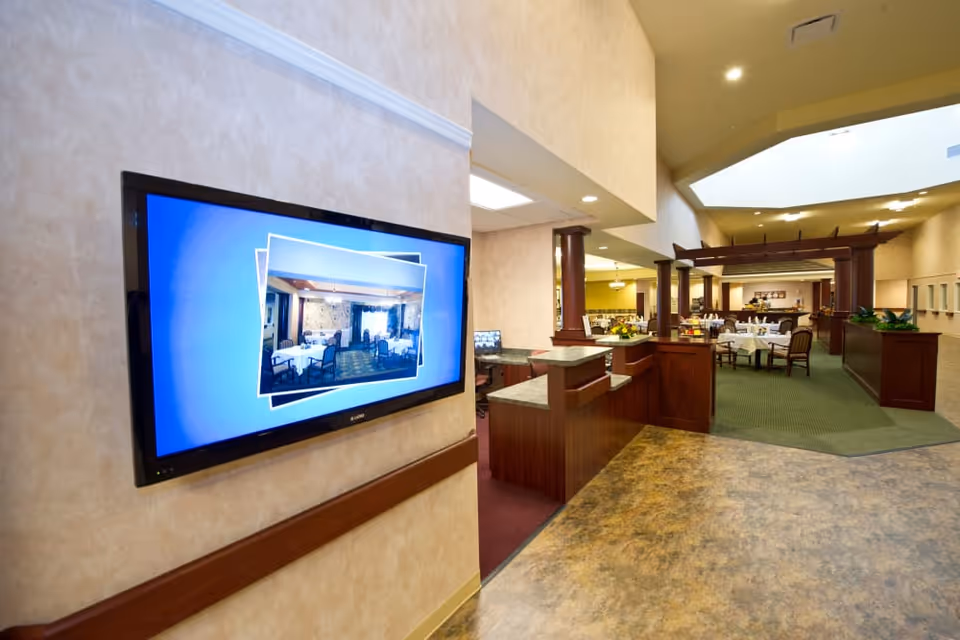 Interior view of a senior living facility dining area with tables and chairs set for meals, visible through an open space with wooden columns and a green carpet. A flat screen TV mounted on the wall displays an image of another dining room.