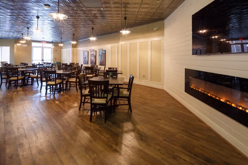 A spacious dining room with multiple wooden tables and chairs arranged neatly on a wooden floor. The room features a modern electric fireplace on the right wall beneath a mounted flat-screen TV. The ceiling has a decorative pattern with hanging light fixtures, and large windows at the far end allow natural light to enter.
