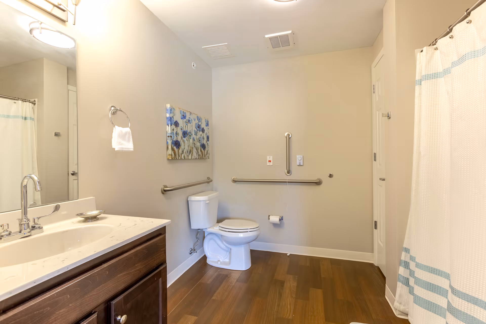A clean and spacious bathroom with a white toilet, a wooden vanity with a white countertop and sink, a large mirror above the sink, a towel ring with a white towel, a shower with a white curtain featuring blue stripes, wooden flooring, and grab bars installed on the walls near the toilet for accessibility. A floral painting is hung on the wall above the toilet.