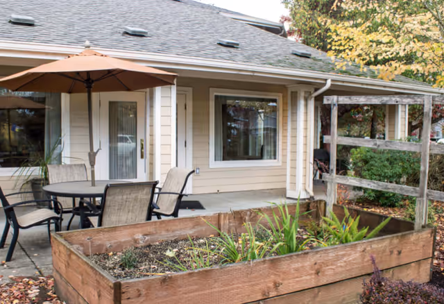 Outdoor patio area at Woodside Senior Living featuring a round table with four chairs and a large umbrella. The patio is adjacent to a beige building with white trim, windows, and a door. In the foreground, there is a raised wooden planter box with some plants and greenery. Trees and shrubs surround the area.