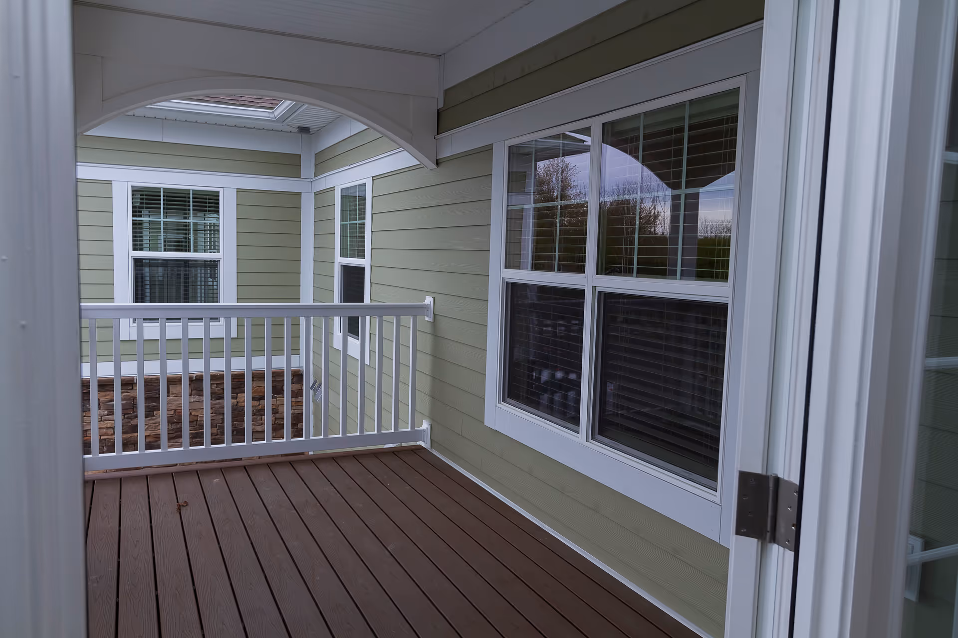 Covered outdoor balcony or porch area with wooden flooring, white railing, and green siding walls with white trim around windows and door.