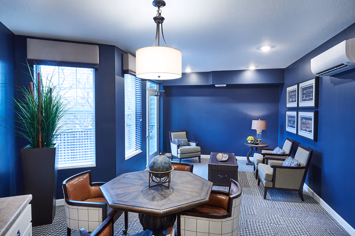 A cozy interior room with dark blue walls featuring a round wooden table surrounded by four cushioned chairs in the foreground. In the background, there are three armchairs with patterned pillows, a small round side table with a lamp and flowers, and framed artwork on the wall. Large windows with blinds allow natural light to enter, and a tall potted plant is placed near the windows. The room has a modern ceiling light fixture and an air conditioning unit mounted on the wall.